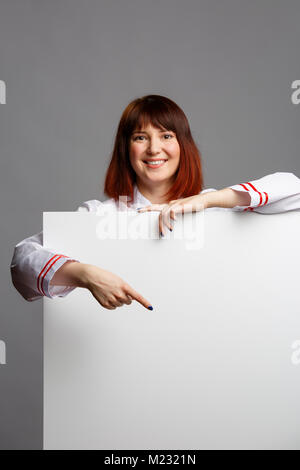 Image of smiling female cook in white robe pointing finger at empty sheet for inscription Stock Photo