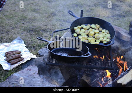 Two cast iron frying pans, sit on a grate over top of a hot fire.  Cooking an early morning breakfast with breakfast sausages and sliced potatoes Stock Photo