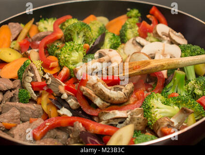Side view of wooden spoon stirring beef and vegetables in cast iron skillet - Stir frying Stock Photo