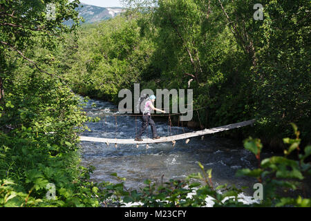 Summer hiking - young woman tourist and traveler with backpack behind her shoulders crossing mountain river on pedestrian suspension bridge in summer Stock Photo