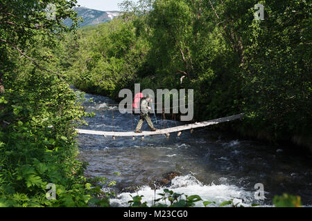 Summer hiking on Kamchatka - woman tourist and traveler with backpack behind her shoulders crossing mountain river in forest on suspension bridge Stock Photo