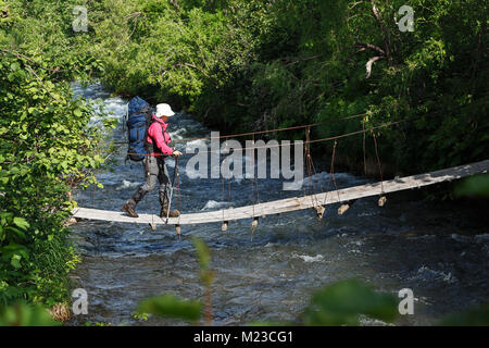 Summer hiking on Kamchatka - young woman hiker with backpack behind her shoulders crossing mountain river on pedestrian suspension bridge in summer Stock Photo