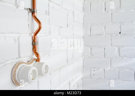Retro socket made of white ceramic with open electrical wiring. Against the background of a white brick wall, electrical wiring in old copper pipes. S Stock Photo