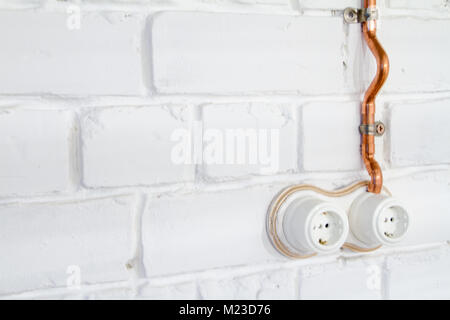 Retro socket made of white ceramic with open electrical wiring. Against the background of a white brick wall, electrical wiring in old copper pipes. S Stock Photo