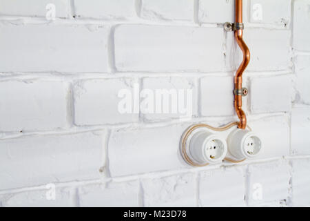 Retro socket made of white ceramic with open electrical wiring. Against the background of a white brick wall, electrical wiring in old copper pipes. S Stock Photo