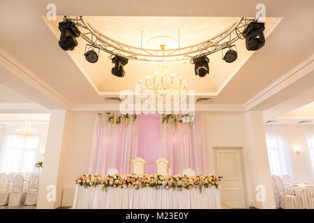 wedding, design, party concept. in the center of the hall there is a table for newly married couple richly decorated with flowers, under this place there is marvelous chandelier and lots of spotlights Stock Photo