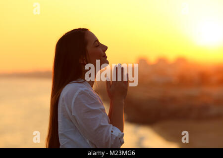 Side view backlight portrait of a woman praying and looking above at sunset Stock Photo