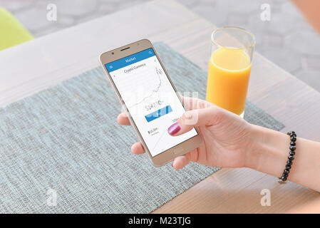Crypto currency graph on smart phone app. Woman holding phone. Juice and table in background. Stock Photo