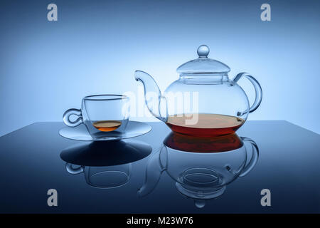 Studio image of transparent glass cup and teapot set with small amount of golden brown black tea at the bottoms of teapot and cup. Set is reflected in Stock Photo