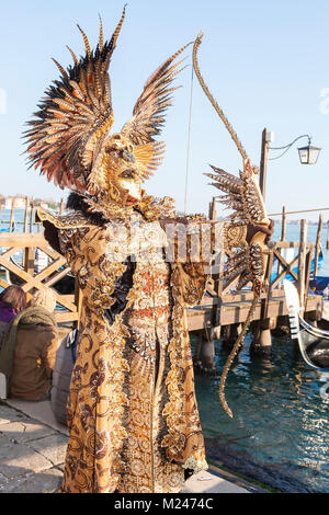 Venice, Veneto, Italy 4th February 2018. Man dresssed as an archer in a spectacular colourful historical costume  and mask with bow posing at the lagoon in Piazzetta San Marco  at the Venice Carnival . Stock Photo