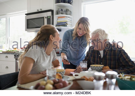Smiling daughters and senior father using smart phone, eating at kitchen table