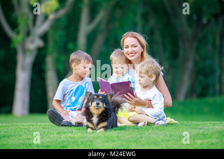 Young mother reading book to her small sons while sitting on grass in park. Children and mother enjoying beautiful summer day. Pet dog lying beside, o Stock Photo