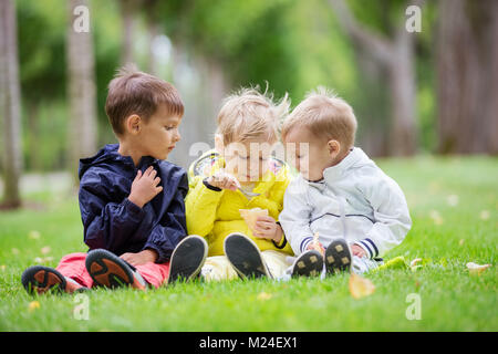 Young boys sitting on grass in  spring park, one brother eating ice cream while the other two watching him Stock Photo