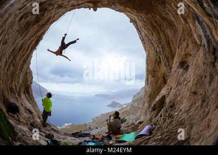 Rock climbers in cave: leading climber swinging on rope after falling of cliff, belayer and female climber watching him Stock Photo