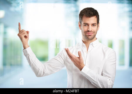 Young man wearing shirt and standing indoor while raising his hand and pointing up something with his finger. Stock Photo