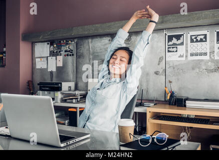 Asian creative designer raise arm up and close eye in front of laptop computer on desk,relax from hard work in office. Stock Photo