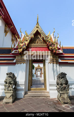 Two statues in front of a decorated and open door and gate at the Wat Pho (Po) temple complex in Bangkok, Thailand, on a sunny day. Stock Photo