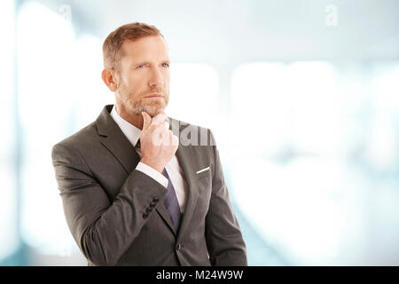 Middle aged businessman wearing suit while standing at the office and seemd deep in thought. Professional man looking away with hands on his chin. Stock Photo