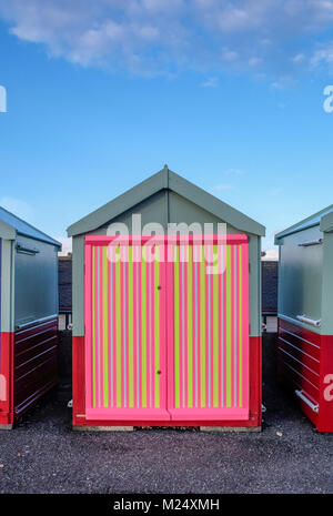Brighton beach hut with a multi coloured door of yellow, pink, black stripes behind is blue sky and infront is the concrete promenade.