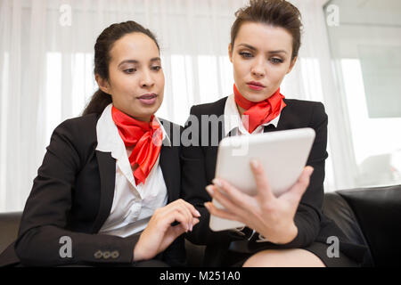 Portrait of two young businesswomen wearing black and red uniform using digital tablet  sitting on sofa in modern office and looking at device screen Stock Photo