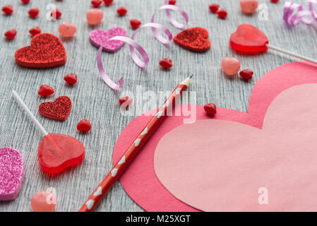 Pink and red heart card lying on table with red pencil and red and pink Valentine's Day candies with grey textured background. Stock Photo