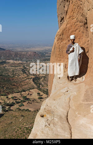 Priest standing on an exposed narrow ledge at the entrance to the rock-hewn church Abuna Yemata, Hawzen, Gheralta Mountains, Tigray, Ethiopia Stock Photo