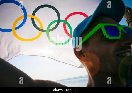 RIO DE JANEIRO - MARCH 10, 2016: Illustrative editorial of smiling Brazilian athlete holding Olympic flag at sunrise view over the beach shore in cele Stock Photo
