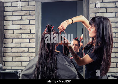 Young female hairdresser cutting and modeling brown hair by scissors and comb of attractive brunette client while she is sitting in armchair in beauty Stock Photo