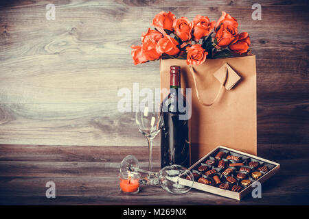 Red wine bottle, box of chocolates, roses in a paper bag on wooden table. Valentines day celebration concept. Copy space. Stock Photo