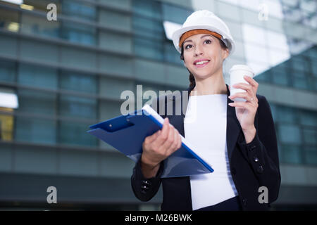 Smiling woman architector in suit and hat is exploring documents with project and drinking coffee near the building. Stock Photo