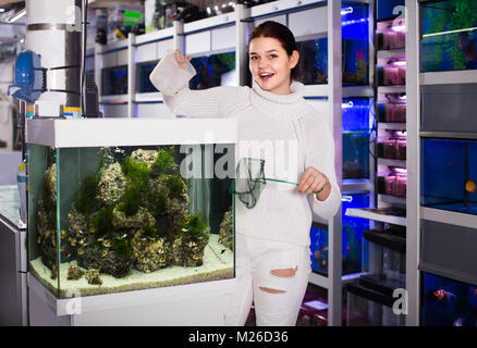 smiling european girl holding aquarian net and water container next to aquarium with colorful fish in aquarium shop Stock Photo