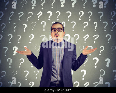 Young elegant in suit shrugging shoulder while looking with lack of knowledge on backdrop with question marks. Stock Photo