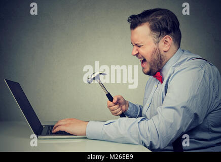 Young angry man frustrated with malfunctioning laptop and holding hammer ready to crush it. Stock Photo