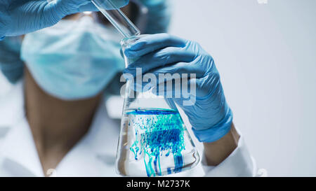 Scientist in gloves pouring blue acid into test tube with transparent liquid Stock Photo