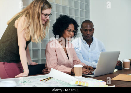 Diverse group of work colleagues working together on a laptop at a desk with blueprints in a modern office Stock Photo