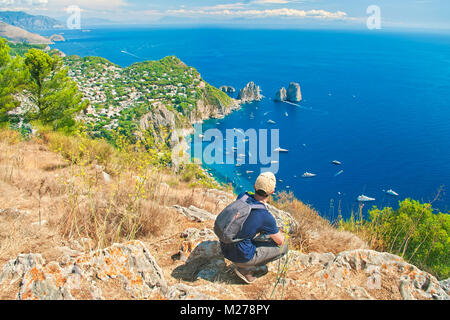 solo traveler resting after climbing up mount Solaro overlooking famous faraglioni rocks on sunny summer day, Capri, Italy Stock Photo