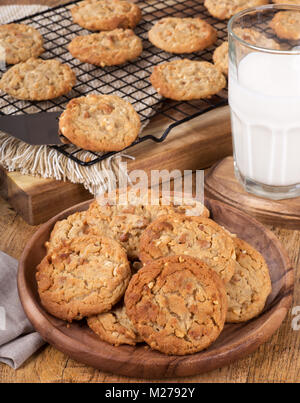 Peanut butter cookies on a wooden plate with cookies on a cooling rack and glass of milk in background Stock Photo