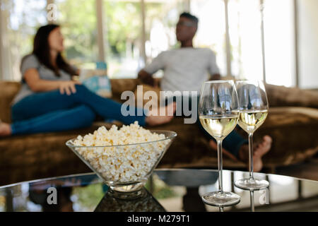 Bowl of popcorn and wine glasses on table in front with couple sitting and relaxing on couch in background. Stock Photo