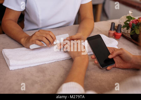 Woman using smart phone while receiving manicure in nail salon. Female client with mobile phone getting manicure and nail care procedure by beautician Stock Photo