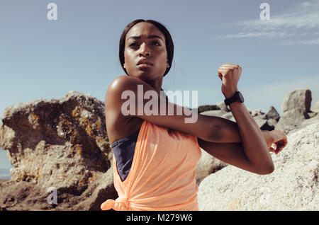 Sportswoman stretching her arm muscle at the beach, looking aside. Fit young african female doing warmup exercise outdoors. Stock Photo