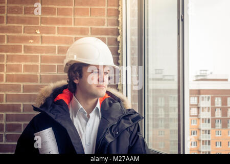 Young architect with white helmet holding blueprint and smiling look in the window. Soft focus, toned. Stock Photo