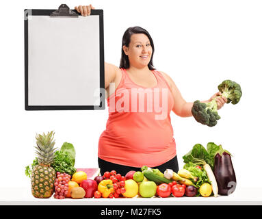 Overweight woman holding a blank clipboard and a broccoli dumbbell behind a table with fruit and vegetables isolated on white background Stock Photo