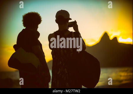 Sunset silhouettes of two unidentifiable tourists taking a photo of two brothers mountains in  Ipanema Stock Photo