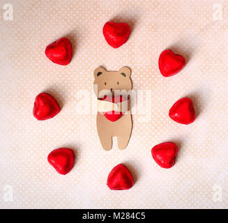 congratulations on the Valentine's Day. A small bear of kraft paper holds in his paws a chocolate candy in the form of a heart in a red foil Stock Photo