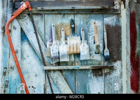 Image of used paint brushes and a saw hung on the door of an old shed with strokes of paint on it. Stock Photo