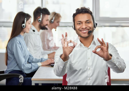 Happy call centre worker giving ok gestures. Portrait of handsome indian black man makes all right, OK, sign using both hands. Everything is fine. Stock Photo