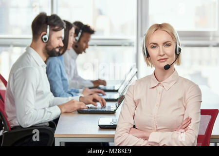 Female serious call center operator with folded arms. Blond mature woman with serious face. Bright windows background. Stock Photo