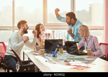 Black male leader reported good news, everyone is happy. Creative people designers extremely happy at messy desk in office. Stock Photo