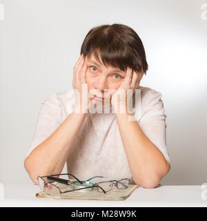 Frustrated woman holds her head in her hands. On the table in front of her is a newspaper, two glasses and a magnifying glass. Poor vision, glasses ar Stock Photo