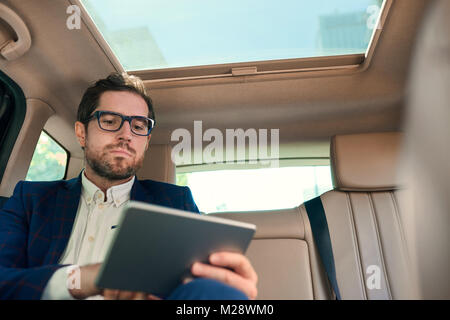 Focused young businessman sitting in the backseat of a car working on a digital tablet while being driven through the city Stock Photo